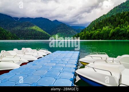 Jetty with white catamarans on the Ritsa Lake in Abkhazia. Mountain lake with green pine forest hills at the shore Stock Photo