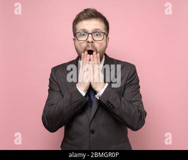 Surprised man with glasses and a suit looks at the camera with big eyes and covers his open mouth with hands. Stock Photo