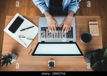 Top view of a millennial woman working at office desk.Human hands typing on laptop. Laptop, diary, eye glasses, smartphone, pen, coffee cup and plants Stock Photo