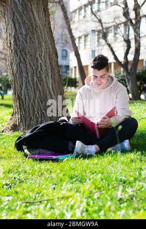 Teenager sitting on the school grass while studying with a book Stock Photo