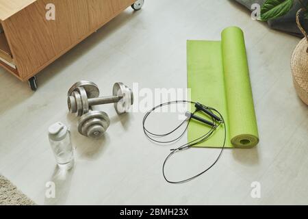 Two metallic dumbbells, skipping-rope, plastic bottle of water and rolled green mat for exercising on the floor of domestic room Stock Photo