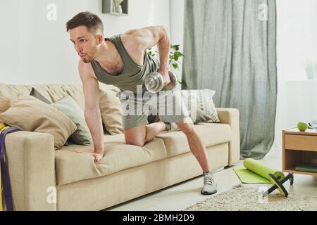 Young sportsman in activewear leaning on couch while doing exercise with dumbbells for arms and biceps during morning workout at home Stock Photo