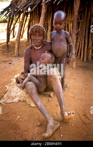 Grandmother Gado sitting on a hide together with her two grandchildren Aike and Zubo(upright) in front of their hut in Kaina. Stock Photo