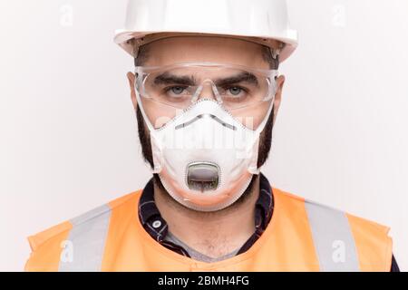 Serious male technician in orange workwear, protective hardhat, eyeglasses and mask looking at you against white background Stock Photo