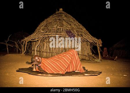 Man rests in the darkness in front of a hut on a hide wih his head on a Hamer stool. Stock Photo