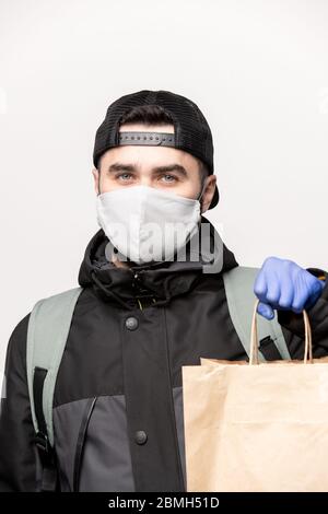 Casual guy in black jacket and cap with backpack holding paperbag with your order from online shop while delivering it during quarantine Stock Photo