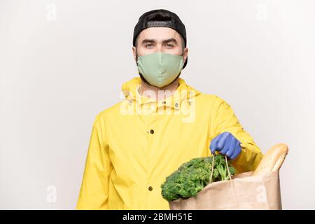 Young man in yellow jacket and black cap with respirator on face and gloves on hands carrying paperbag with fresh vegetables from supermarket Stock Photo