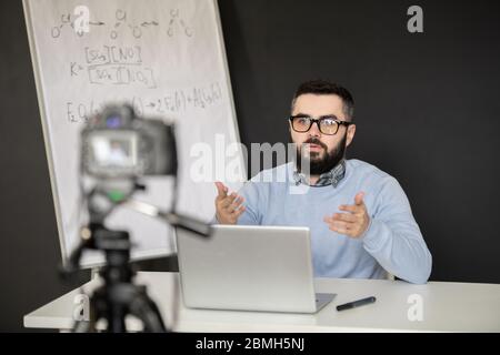 Contemporary school teacher in pullover and eyeglasses explaining something to his students during remote lesson of chemistry Stock Photo