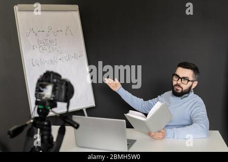 Young confident teacher of chemistry explaining his students formulas on whiteboard while looking at laptop display during online lesson Stock Photo