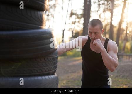 Man fighter training boxing outdoor tire DIY handmade gym in forest fitness workout. Young adult  training right hand hit nature without boxing gloves Stock Photo