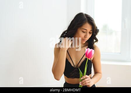 A portrait of charming dark haired young woman with many beautiful pink tulips on the white background near the window. Stock Photo