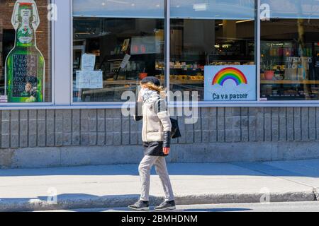 Person wearing a protective mask and a scarf walking in front of a store with a covid 19 rainbow of hope poster on it (the poster says in French: It will pass) , Montreal Canada Stock Photo