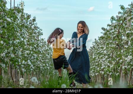 Happy beautiful mom and daughter in elegant clothes dancing among the blooming garden Stock Photo