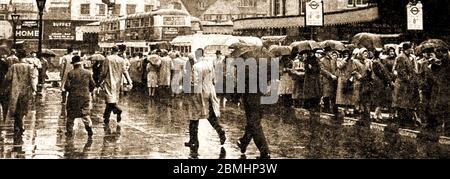 WEATHER - RAINY BRITAIN -Londoners in the rain. - 1950's street scene with passengers waiting in queues at a bus stop wearing headscarves, raincoats & carrying umbrellas. Stock Photo