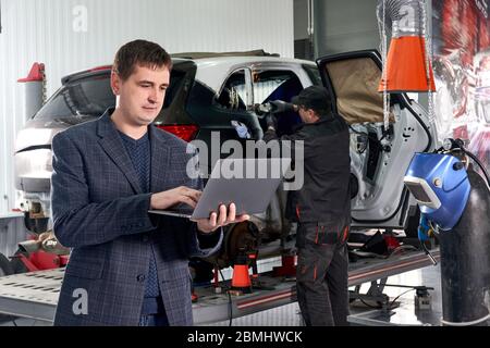 Manager holding laptop while standing near car in auto repair shop with mechanic repairing car on background Stock Photo