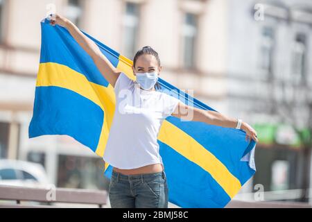 Beautiful smiling Swedish girl holds a flag while wearing a protective face mask outdoors. Stock Photo
