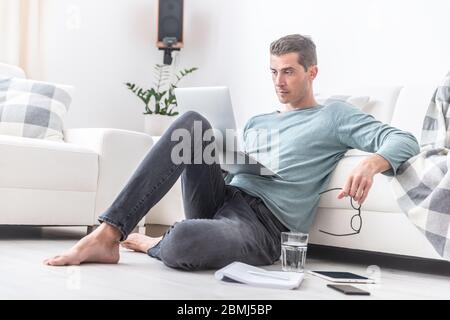 Male freelancer sits barefoot on the living room floor working on a laptop. Stock Photo