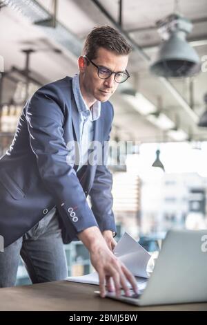 Casually dressed businessman standing in front of a coffee house table touching a keyboard of a notebook in front of him. Stock Photo