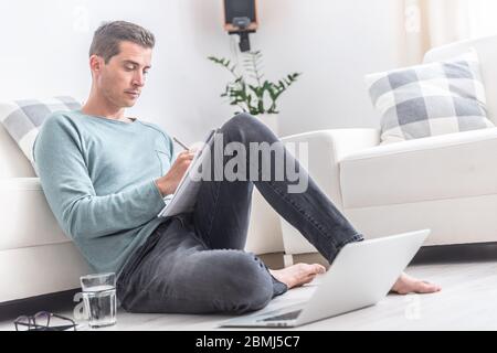 Man working from home writes down on paper while sitting on the floor. Stock Photo