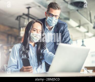 Good looking female and male coworkers look at the computer screen together, wearing face masks. Stock Photo