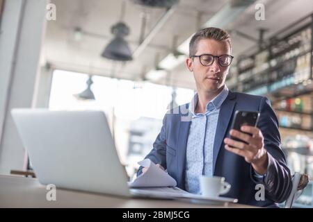 Young businessman types on his mobile phone with a laptop and coffee in front of him in a coffee shop. Stock Photo