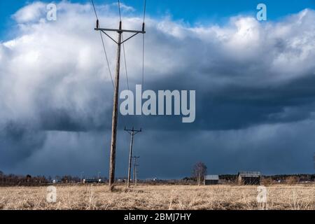 Row of wooden electricity poles with three wires and ceramic isolators in field. Heavy dark blue clouds before thunderstorm, brown faded grass after w Stock Photo
