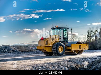 Eye catching yellow road roller with enclosed climate controlled cabin stands on not ready new road, stones, blue sky, clouds, back left side view. Cl Stock Photo