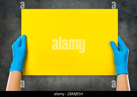Empty yellow card in doctor’s hands in blue gloves with copy space on grunge grey background wall. Stock Photo