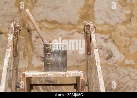 A single paint brush sitting in an old canned fruit tin covered in paint and resting on an old painters ladder in morning sunlight Stock Photo