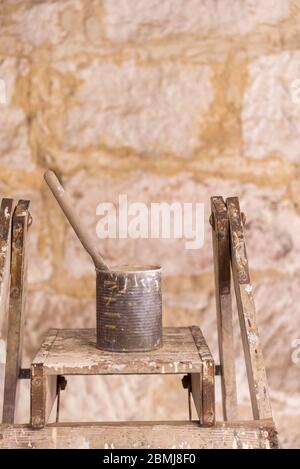 A single paint brush sitting in an old canned fruit tin covered in paint and resting on an old painters ladder in morning sunlight Stock Photo