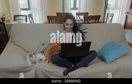 Brunette woman teleworking at home on a sofa with her cat next to her Stock Photo