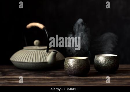 Teapot and tea cups with steaming fresh tea. Traditional japanese chinese iron teapot and ceramic teacups, asian tea set on dark background, copy spac Stock Photo