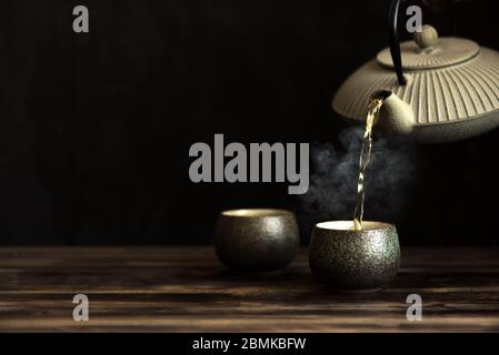 Teapot pouring tea into cups. Traditional japanese chinese iron teapot and ceramic teacups, asian tea set on dark background, copy space. Stock Photo