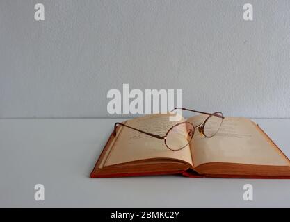 Closeup and selective focus on one lens of eyeglasses placed on open vintage book, red hardcover on white table against white cement wall background Stock Photo
