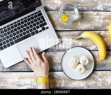 Woman working from home and eating banana and sweet homemade Zephyr or Marshmallow from banana near laptop on wooden table top view.  Time for break Stock Photo