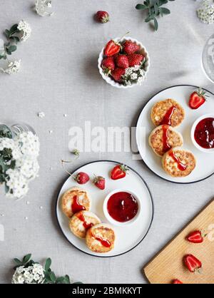 Homemade Ukrainian cheese pancakes Syrniki with red strawberry jam and berries near white flowers on table cloth top view