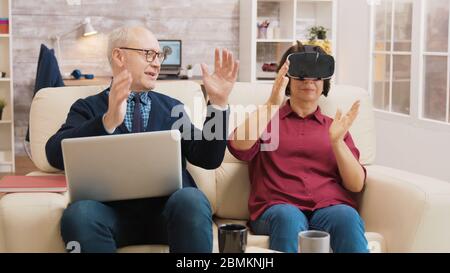Amazed senior woman while using virtual reality goggles on sofa with her husband next to her using laptop. Stock Photo