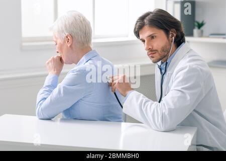 Serious doctor listening to coughing senior patient at clinic Stock Photo