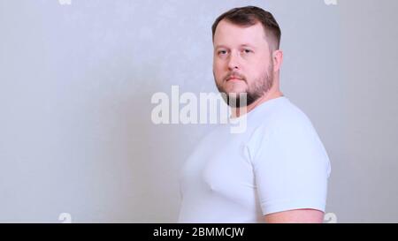 Closeup portrait of a Caucasian big fat man with green eyes looking directly at the camera, wearing a white T-shirt, skeptical suspicious expression Stock Photo