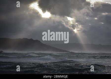 Big waves break on the coast, in the sunlit sea, passing through the clouds at sunset. Galicia, Spain. Stock Photo