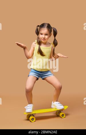Active young girl in yellow shirt and jeans shorts on skateboard over beige background Stock Photo