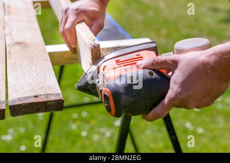 Unrecognizable man in the garden sanding wooden planks. DIY home improvement, restoration, carpentry concept. Hand detail shot. Stock Photo