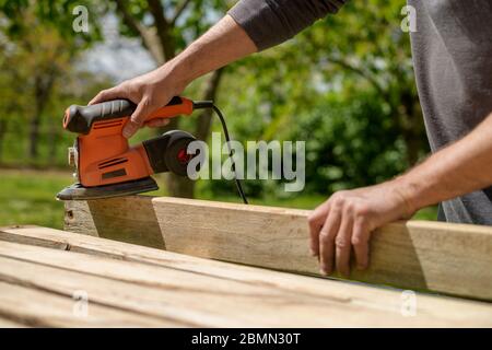 Unrecognizable man in the garden sanding wooden planks. DIY home improvement, restoration, carpentry concept. Midsection hand detail shot. Stock Photo