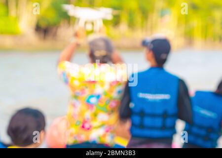 Blurred image of young man in blue and yellow life vest controlling a drone to take pictures of ocean coast with tourists while traveling on a raft in Stock Photo
