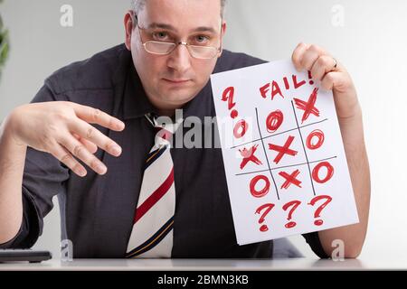 Despondent man holding up a failed tic-tac-toe or noughts and crosses game showing the hand drawn grid with no winner and question marks and text - Fa Stock Photo