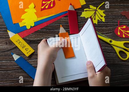 Child holds multi-colored bookmarks origami pencil made of paper new school year on a wooden table. Welcome to school. Children's art project, needlew Stock Photo