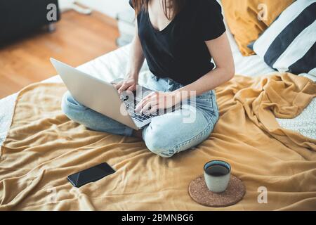 Casual woman working on a laptop sitting on the bed in the house. Female relaxing and drinking cup of hot coffee or tea using laptop computer. Stock Photo