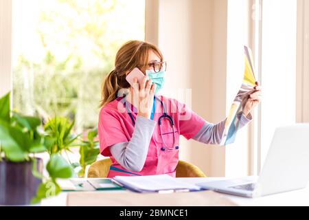 Shot of female doctor wearing face mask while holding x-ray image in her hand and talking on mobile phone. Stock Photo