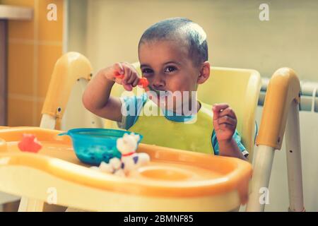 The boy 2 years eats porridge. Children's table. The concept of the child's independence. funny kid in a baby seat. toned Stock Photo