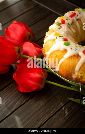 Easter yeast cake or babka covered with icing and decorated with colorful decoration on a dark wooden table and bright tulips, vertical image Stock Photo